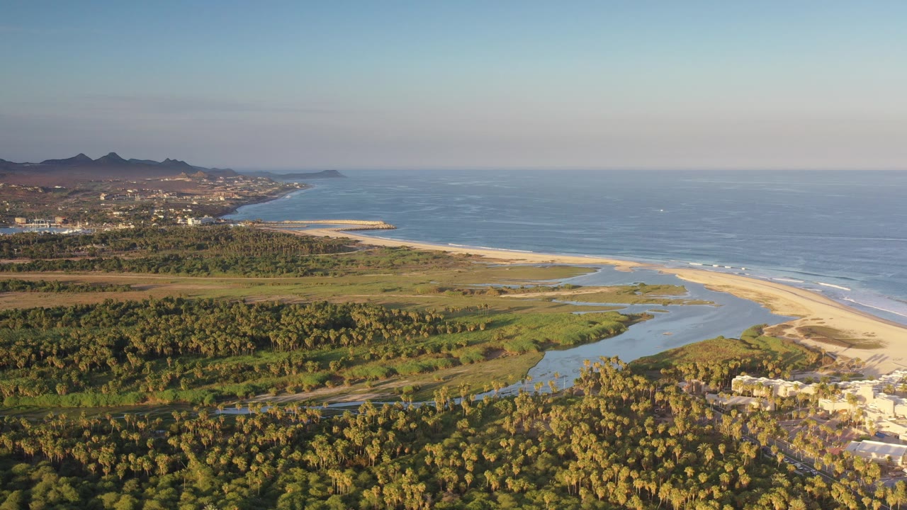 Opening aerial frame over the Los Cabos coast in Mexico, with palm-covered foreground, a lagoon cutting toward a pale sandbar, blue ocean on the right, distant hills on the left, and no visible people.