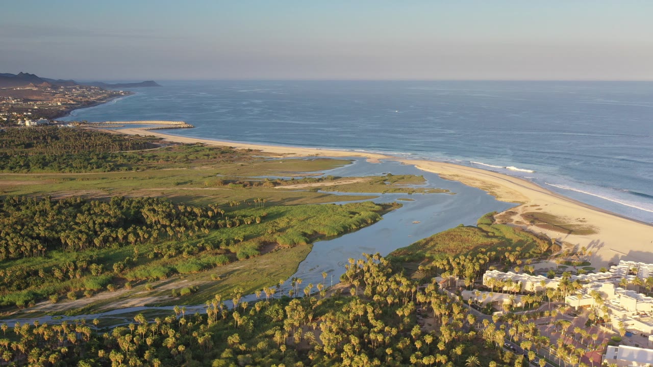 Wide aerial view in Los Cabos, Mexico, showing a winding lagoon behind a sandbar, blue ocean beyond, palms in the foreground, distant shoreline development, and no visible people.