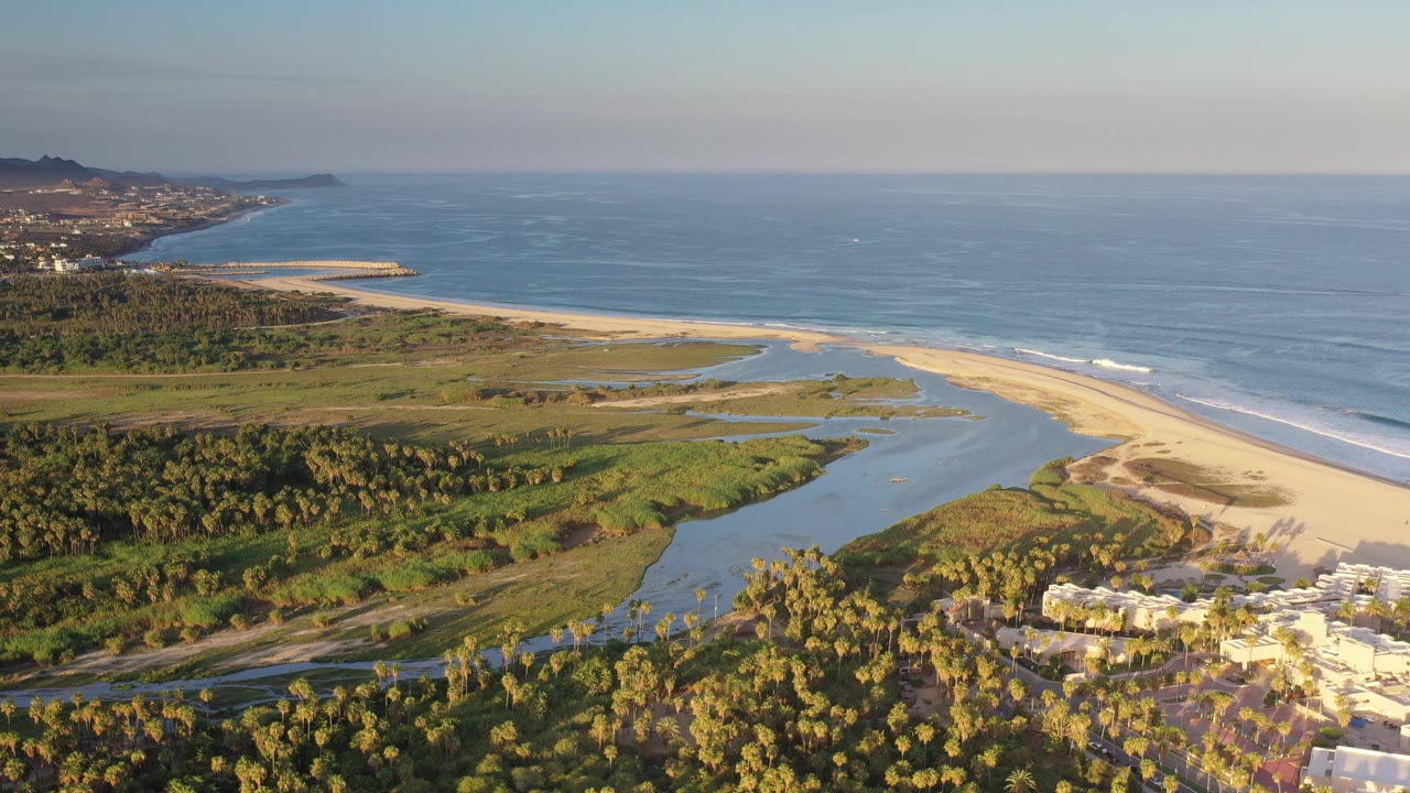 Aerial frame over Los Cabos, Mexico, with the lagoon channel centered, the pale beach curving along the ocean, sunlit palms below, and no visible people.