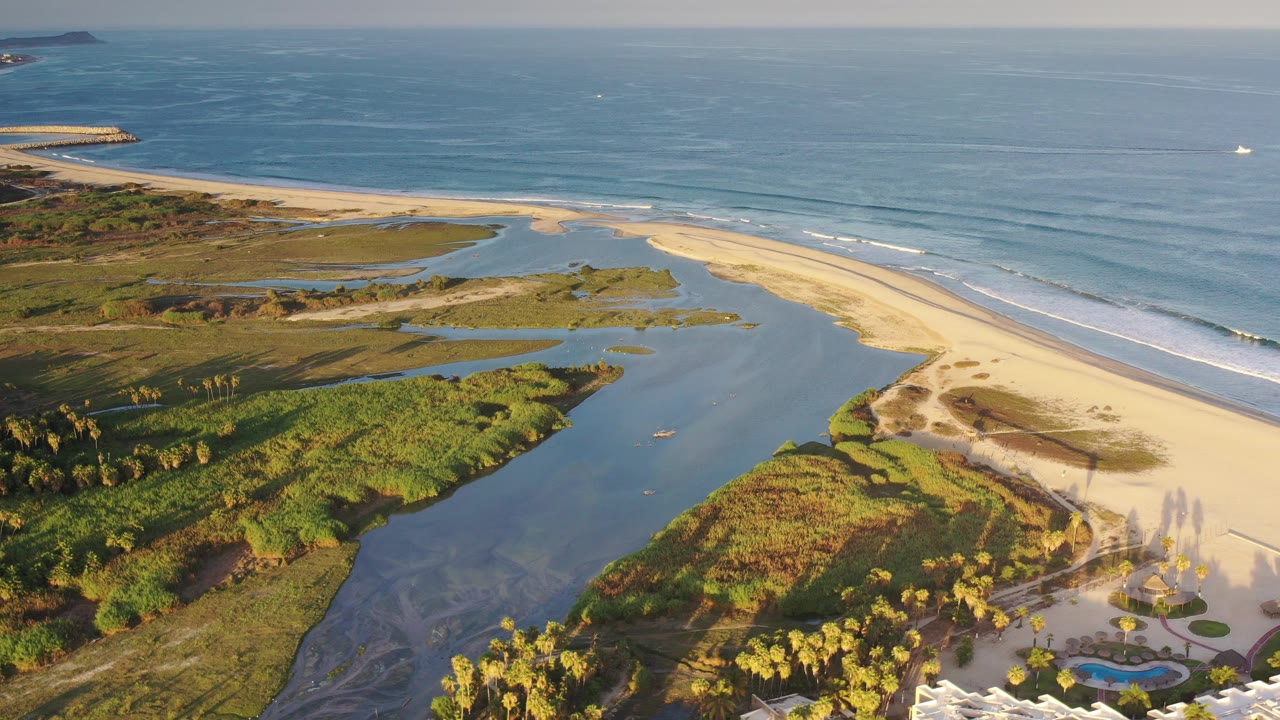 Aerial view over a Los Cabos beach with a curved lagoon, green coastal vegetation, pale sand, blue ocean, small offshore boats, and no visible people.