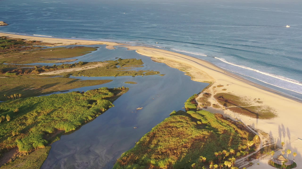 High aerial view in Los Cabos, Mexico, showing blue estuary water, green wetlands, pale beach, rolling ocean waves, and no visible people at the start of the clip.