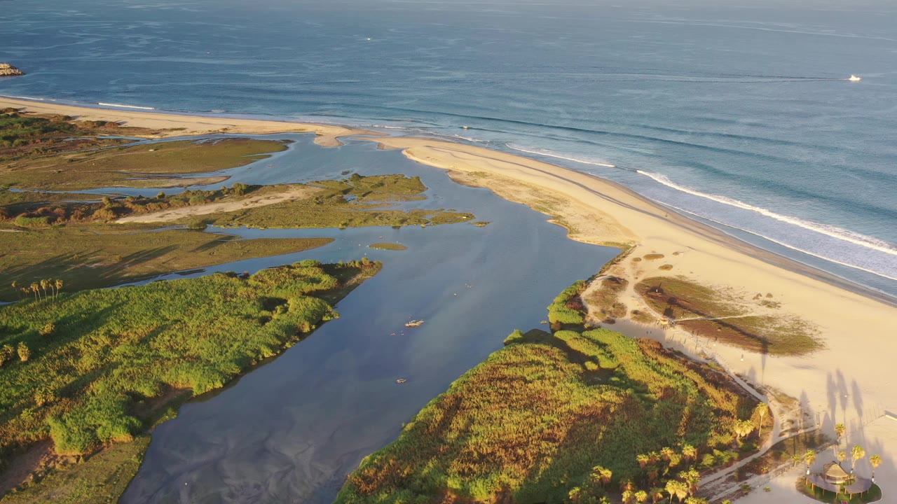 Aerial frame in Los Cabos, Mexico, with the estuary channel centered, green marsh plants in the foreground, sandy shoreline to the right, surf beyond it, and no visible people.