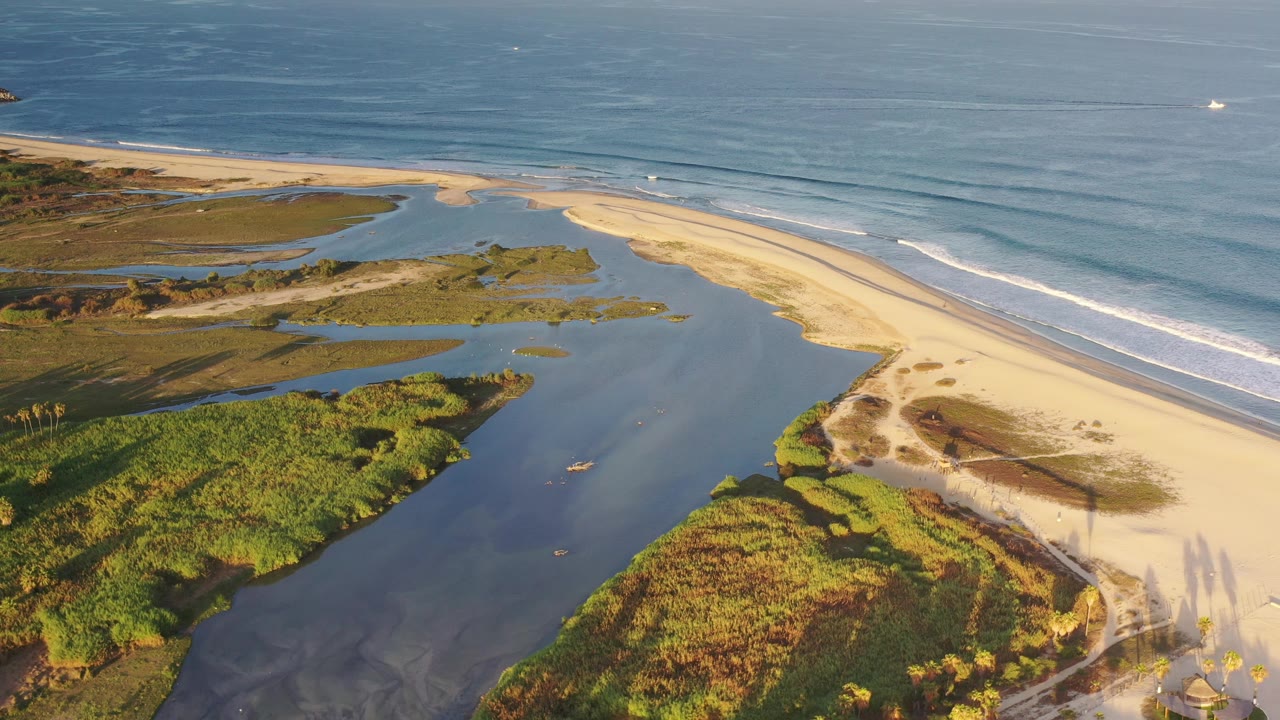 Wide coastal aerial with lagoon water, marsh vegetation, beach, and waves.