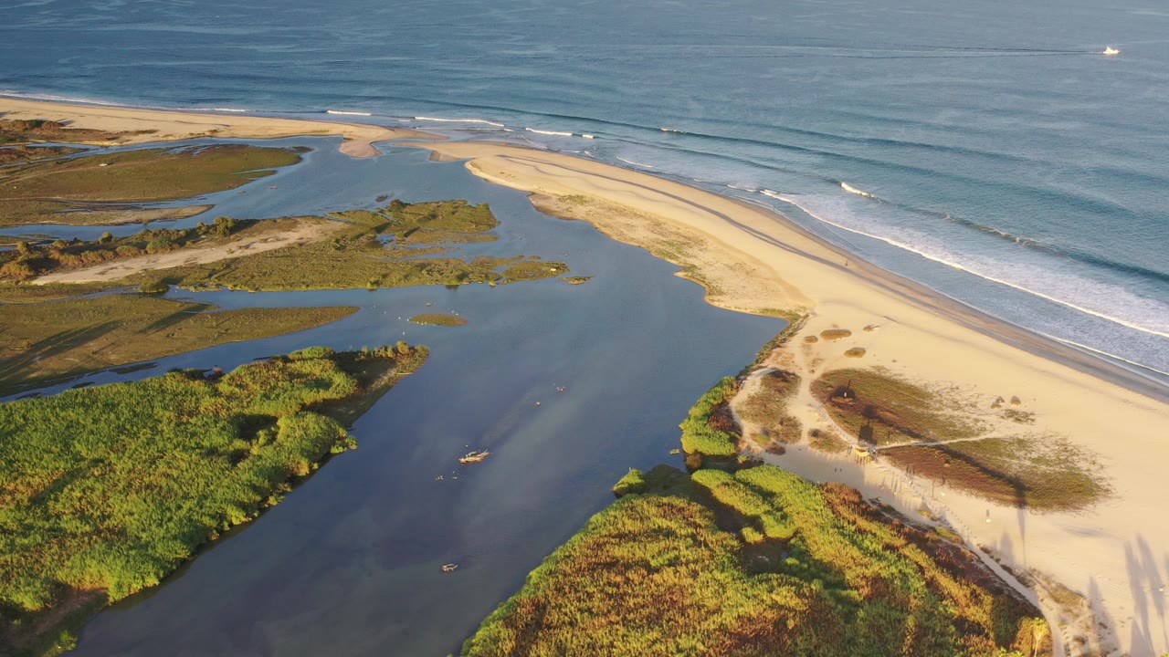 Aerial frame over the Los Cabos coastline shows blue ocean waves beside a pale sandy beach, dark lagoon water, green marsh patches, small boats, and no visible people.