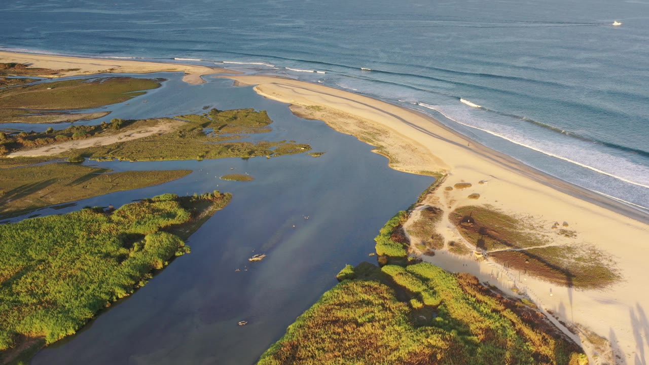 Drone-style aerial view of surf lines, sandbar, lagoon, and green coastal vegetation.