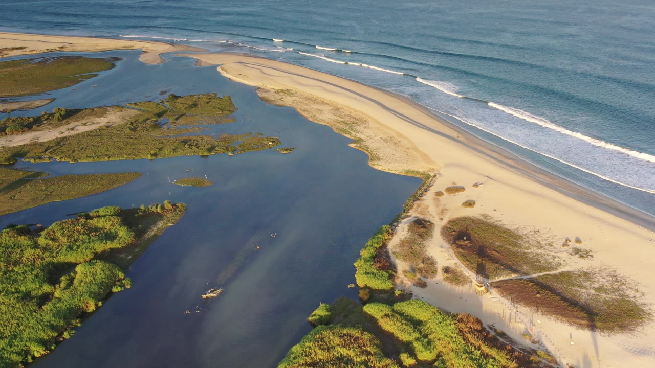 Aerial view in Los Cabos, Mexico, showing a pale sandbar between blue ocean waves and darker lagoon water, with green vegetation at the edge and no visible people.