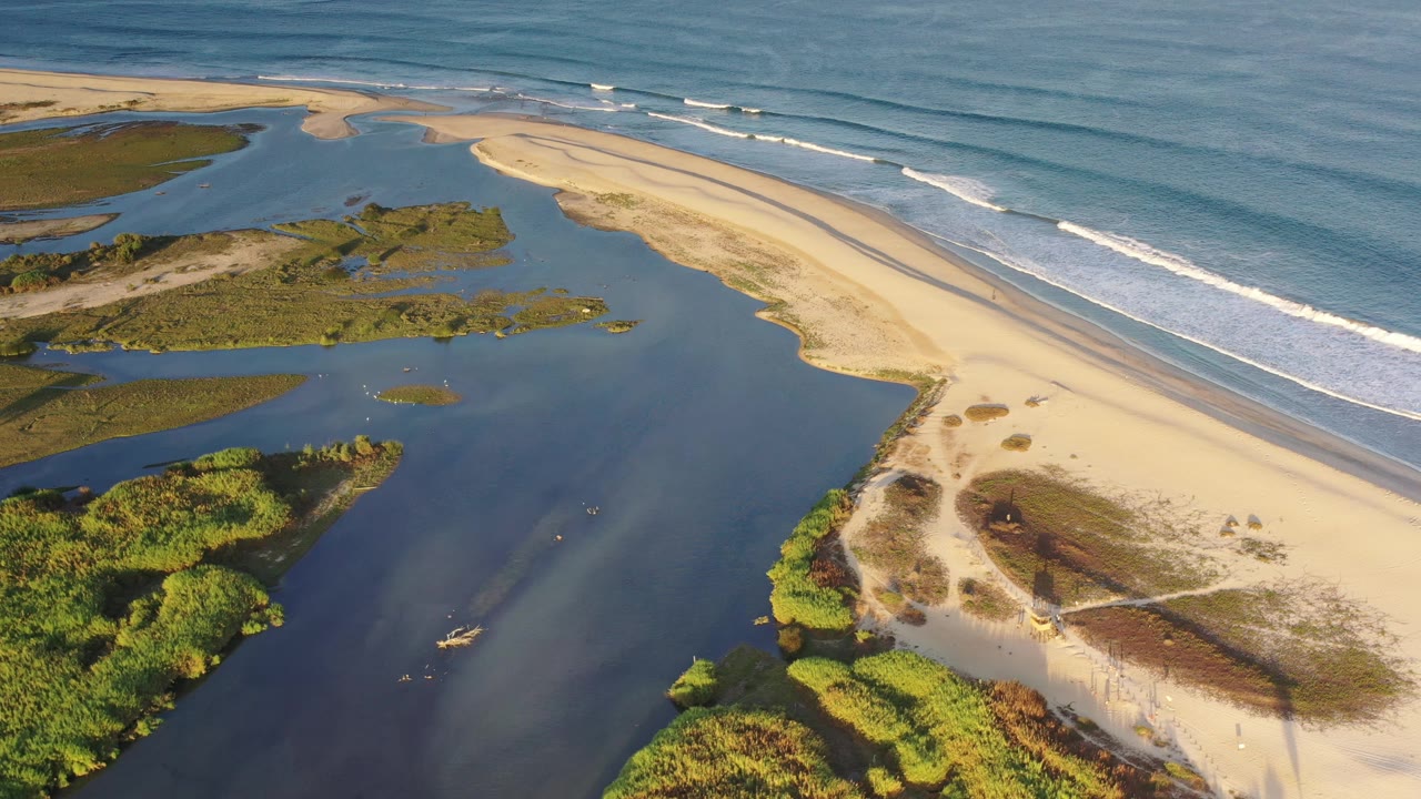 High aerial frame over Los Cabos, Mexico, with the lagoon filling the foreground, a bright sandbar crossing the middle, and parallel wave lines breaking offshore, with no visible people.