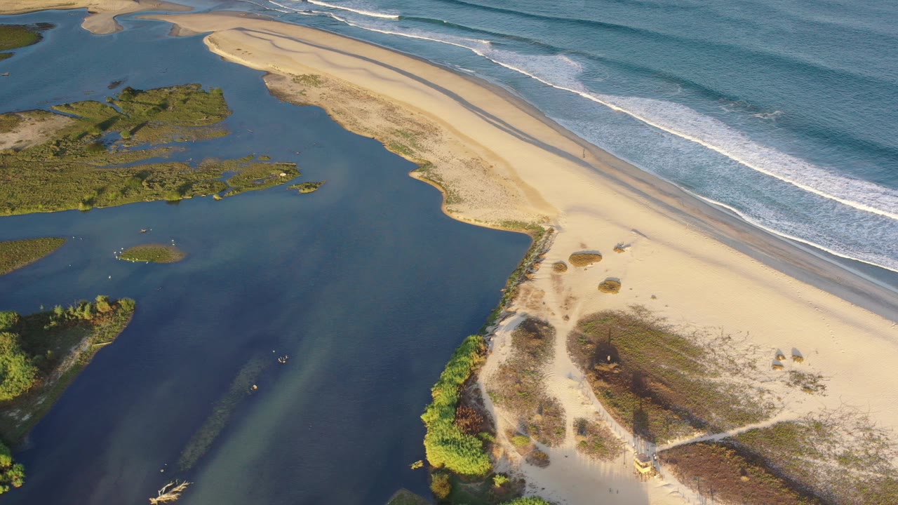 Aerial view in Los Cabos, Mexico showing dark inland water beside pale sand, a narrow sandbar, green coastal plants, small white surf lines, and no visible people.