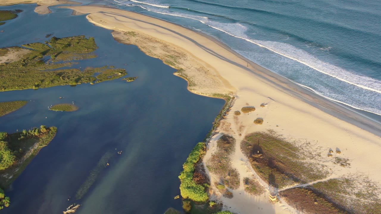 Los Cabos aerial shoreline frame with inland blue water on the left, pale beach and sandbar through the center, ocean waves at upper right, and no visible people.