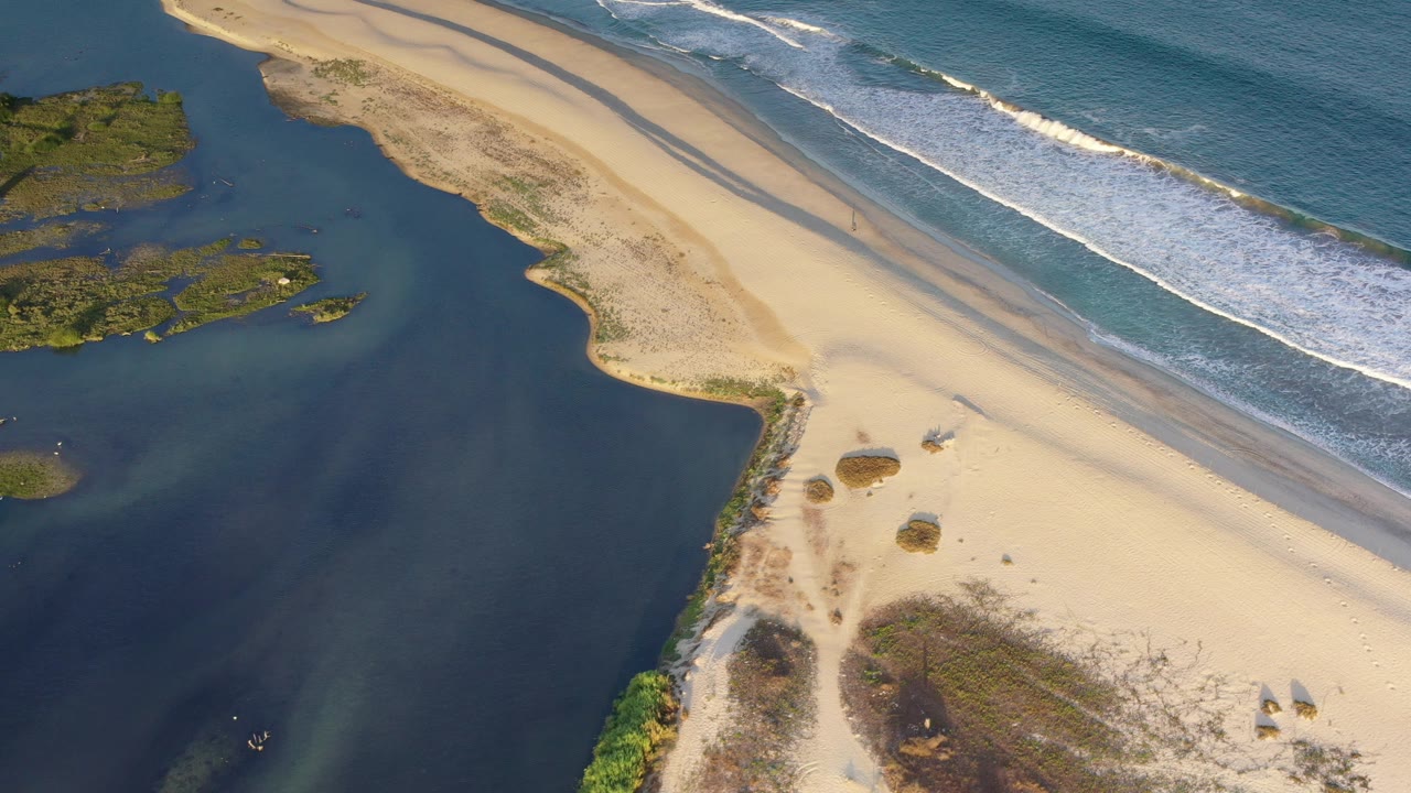Overhead view in Los Cabos, Mexico showing dark estuary water beside a pale sandbar and white ocean waves, with no visible people.