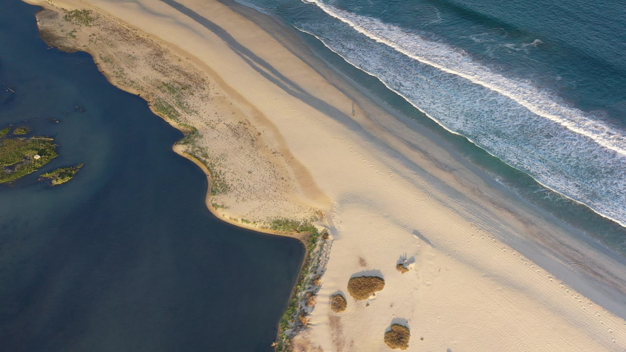 Aerial view over Los Cabos, Mexico, with a pale sandbar between dark lagoon water and blue Pacific surf, plus small beach shelters near the lower edge and no visible people.
