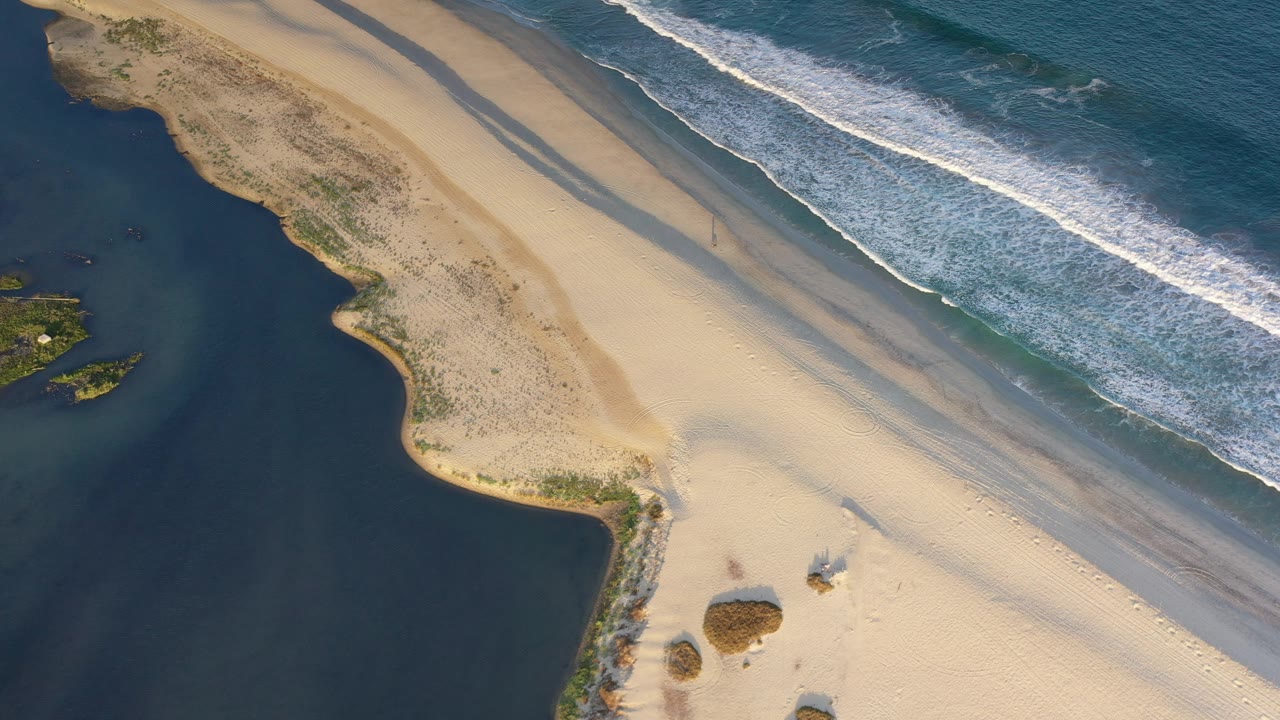 Sand peninsula bordered by dark inlet water and ocean waves.