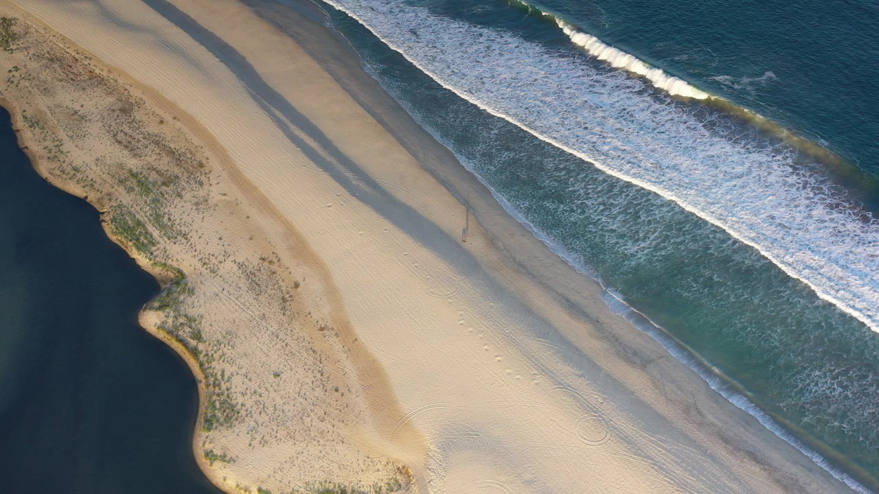 Overhead view in Los Cabos, Mexico, with pale beach sand between white surf and a dark water edge, no visible people.