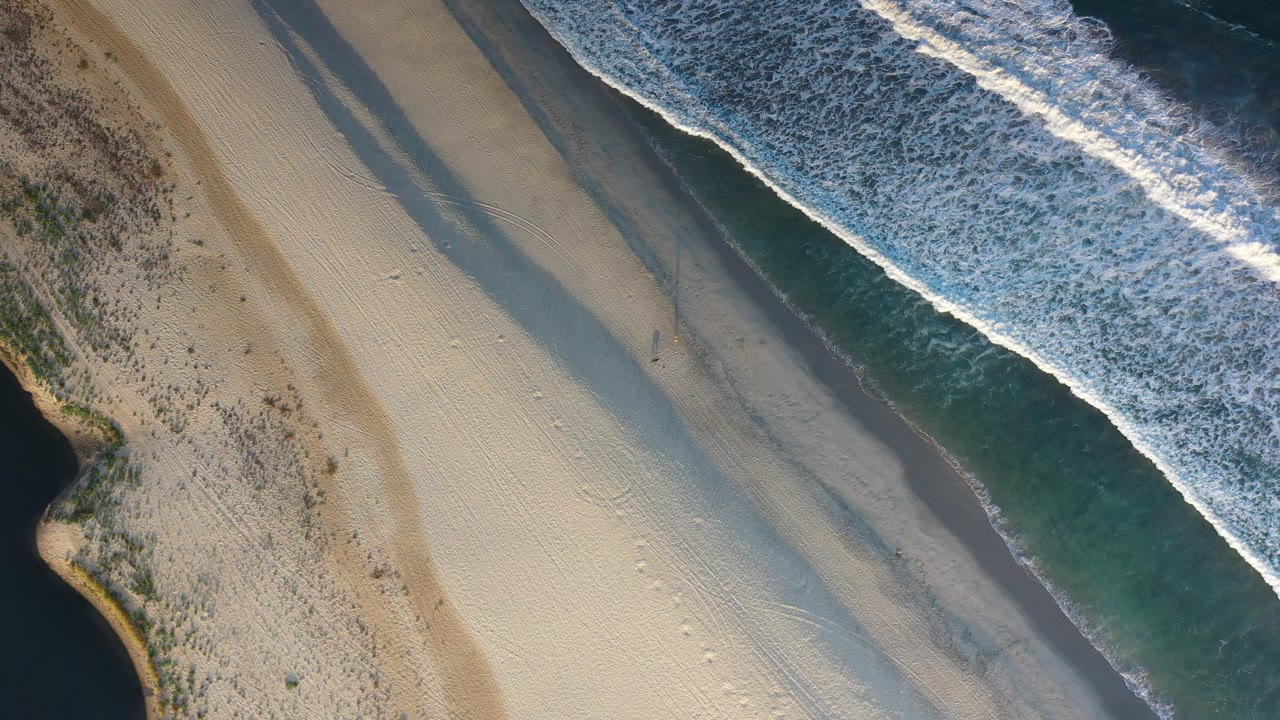 Aerial Los Cabos shoreline frame with a diagonal white surf line, green water, broad gold sand, faint track marks, and no visible people.