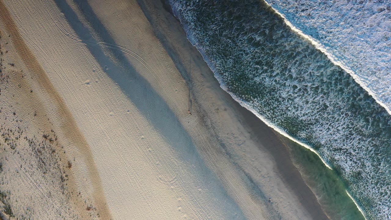 Overhead Los Cabos beach view with dark blue green water and white foam cutting diagonally across pale sand, with no visible people.
