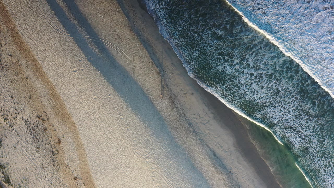Overhead Los Cabos shoreline with a bright foam edge, darker wet sand, pale dry sand, faint tracks, and no visible people.