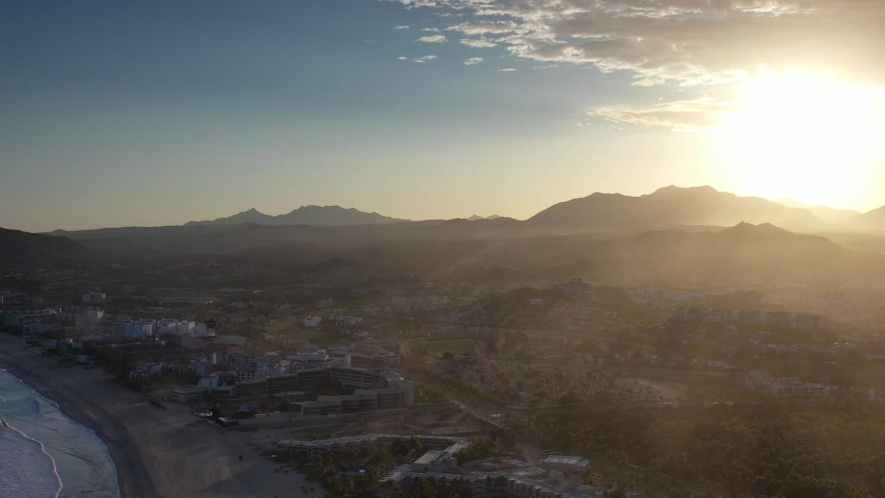 Los Cabos, Mexico aerial frame with ocean surf at left, low buildings below, mountain silhouettes across the horizon, strong sunrise glare at upper right, and no visible people.