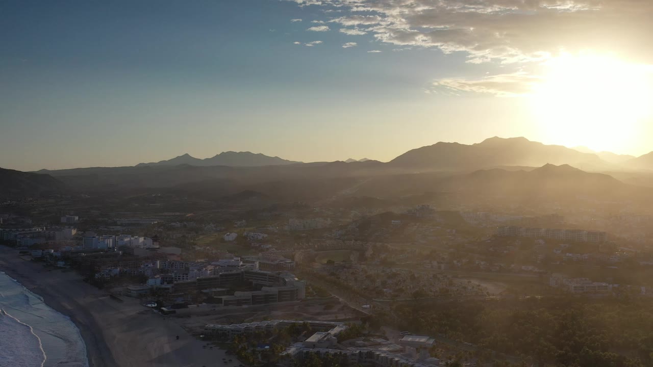 Los Cabos, Mexico aerial view showing the beach curving along the left, a cluster of low coastal buildings below, hazy inland terrain, mountain ridges, and no visible people.