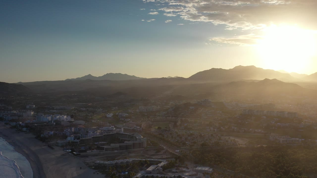 Aerial view of beach, buildings, countryside, and mountains under low sun.
