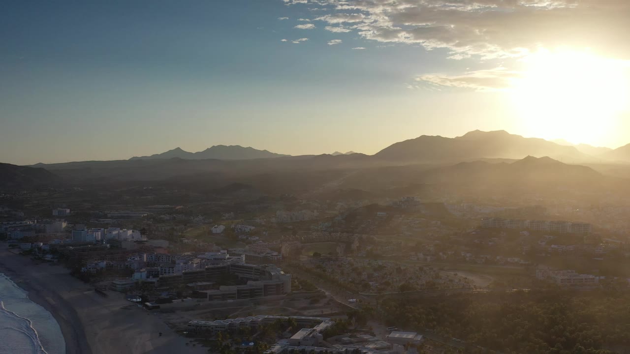 The Los Cabos aerial view holds on the coastal edge and low buildings, with dark open land leading to a mountain ridge and bright sunlight washing the upper right, no visible people.