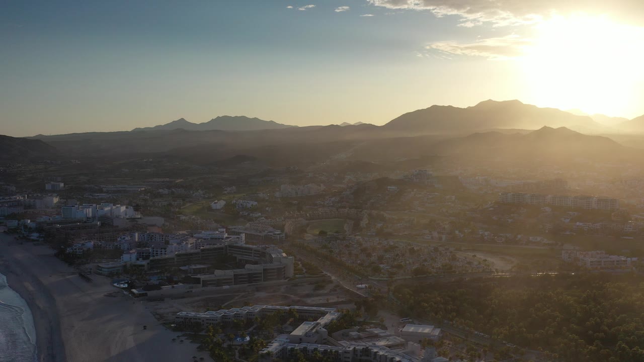 Aerial view of coastal buildings, trees, beach, and mountains at sunrise.