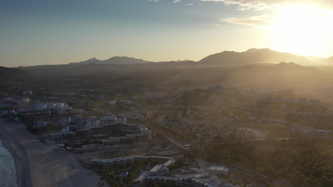 Wide aerial of a beachside town below hazy mountains at dawn.