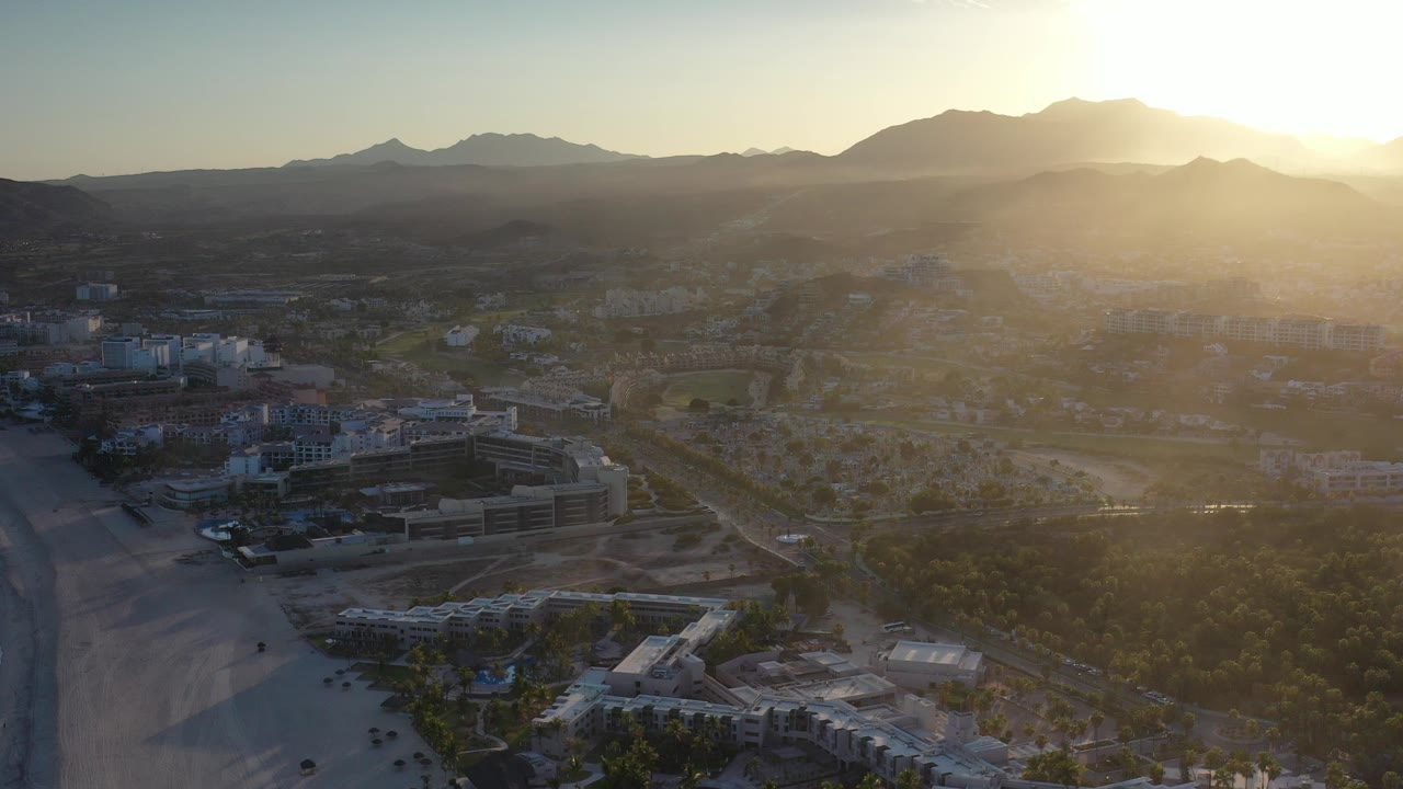 A wide aerial view over Los Cabos, Mexico, shows beachfront buildings, pale sand, roads, trees, and distant mountains under low golden light, with no visible people.