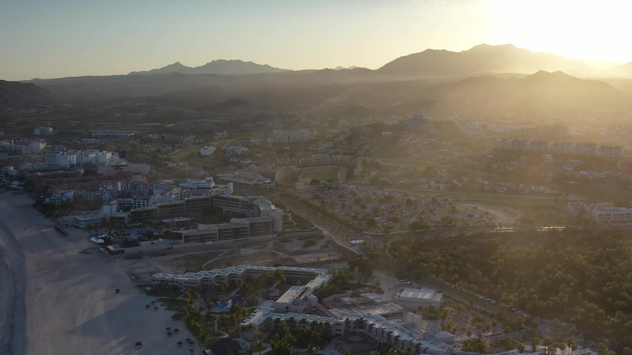 The aerial frame looks across Los Cabos from beachside structures and open sand toward a developed inland area and hazy mountain ridges, with no visible people.