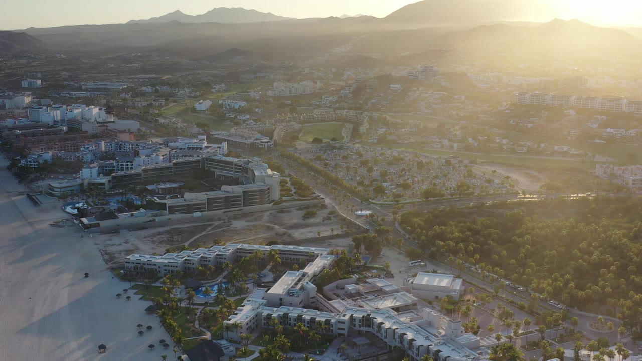 High aerial view over Los Cabos, Mexico, showing beachfront buildings, sandy open ground, tree cover, city blocks, distant mountains, and no visible people.