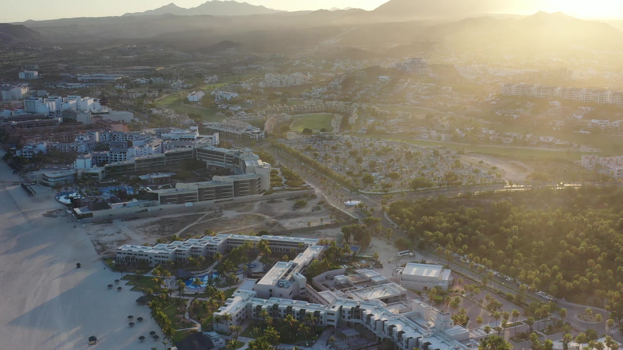 Aerial cityscape beside a beach with bright sun flare and mountains behind.