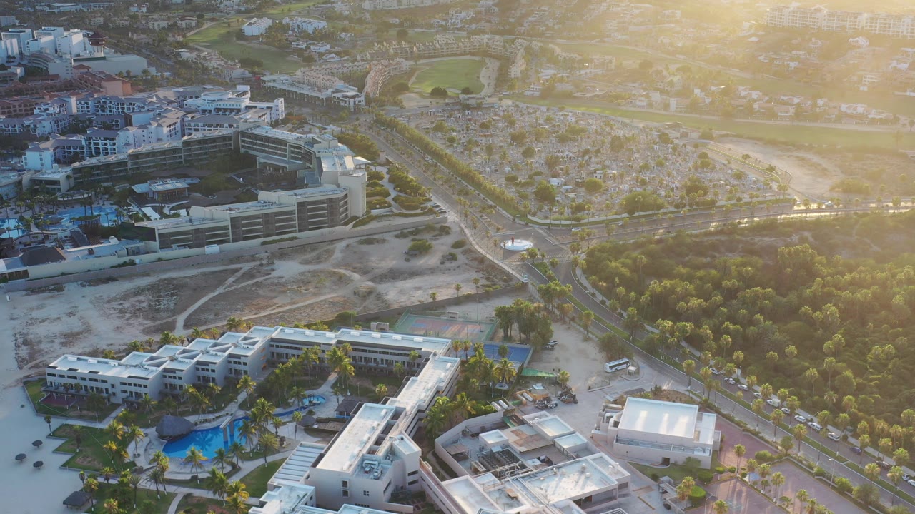 Opening aerial frame over Los Cabos, Mexico, showing white low-rise buildings, a blue pool, roads, trees, dry lots, distant hills, and no visible people.