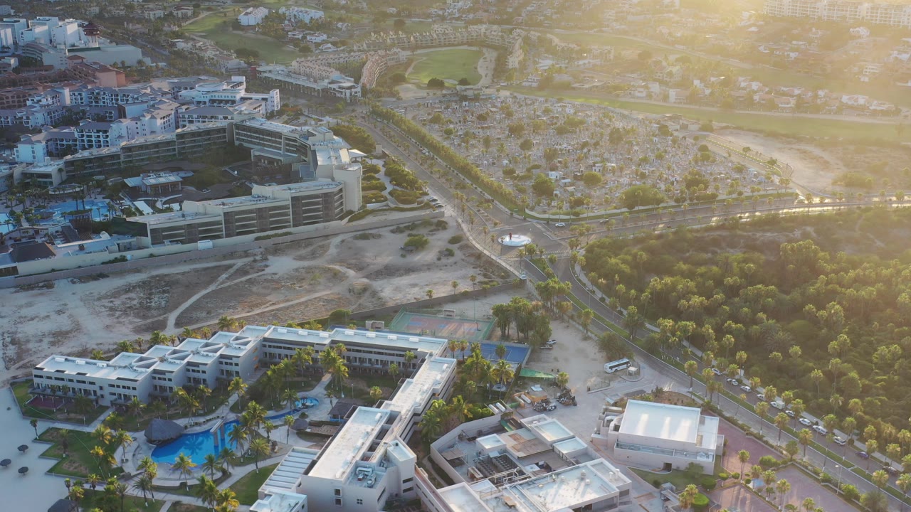 Drone view across white buildings, blue pool, roads, trees, and hazy hills.
