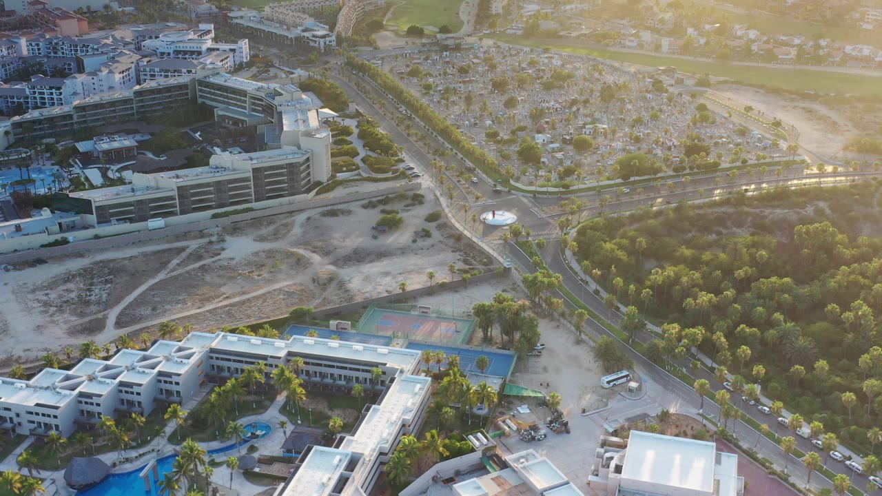 One second into the Los Cabos aerial, the road remains centered with trees on the right, resort-style buildings and pools along the lower edge, and no visible people.