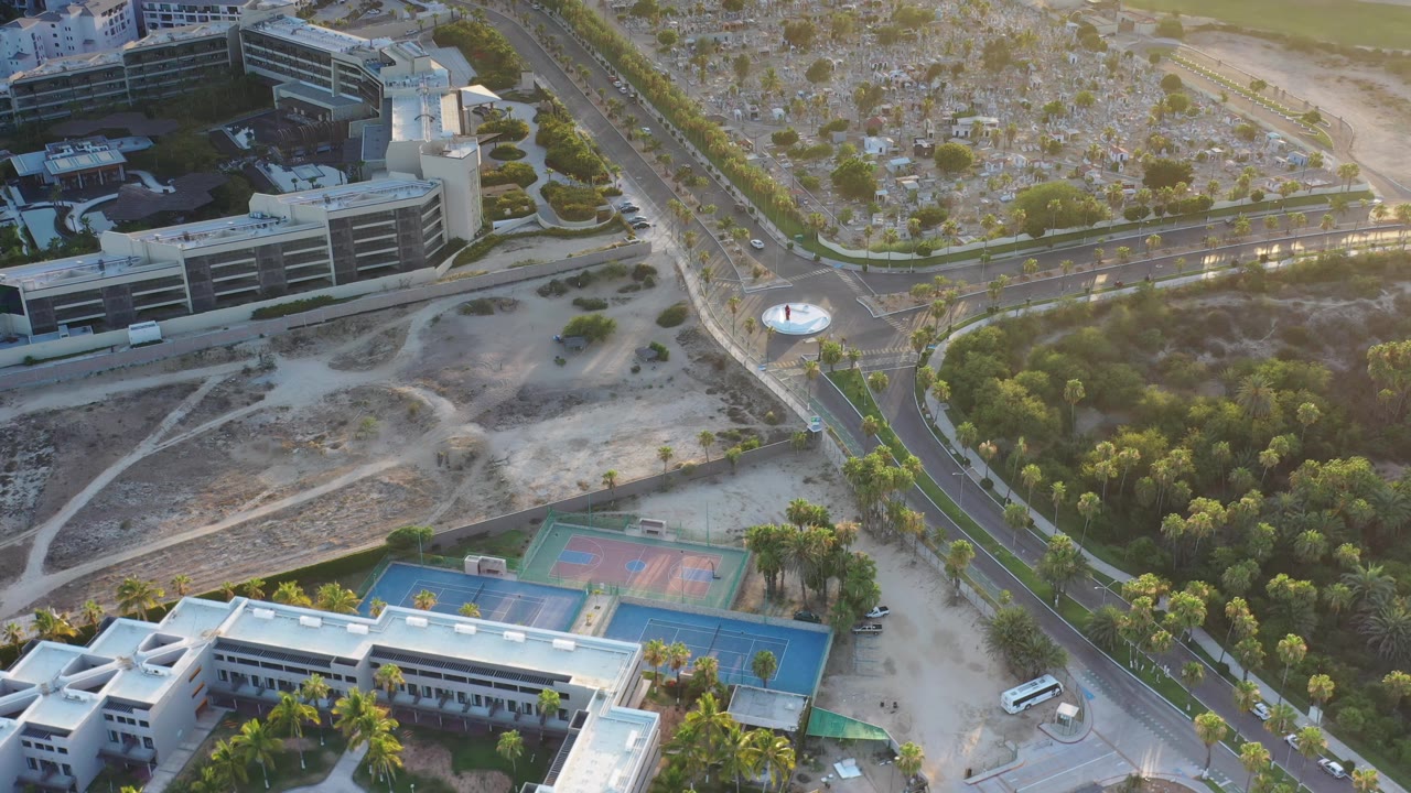 High angled view over Los Cabos, Mexico, with a dark building near the upper left, dry lots, roadside greenery, sports courts, and no visible people.