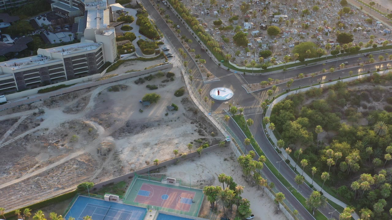 Opening aerial frame over Los Cabos, Mexico, showing intersecting roads, a multi-story building, blue and red sports courts, palms, dry ground, and no visible people.