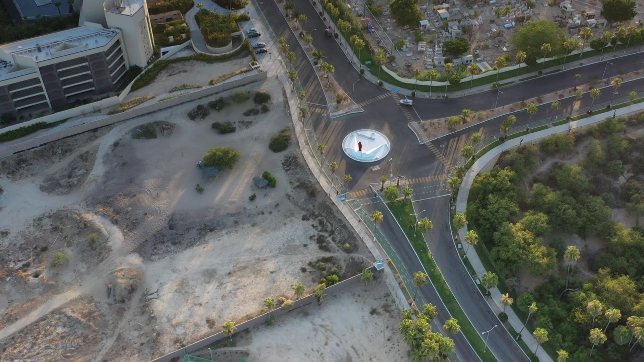 Opening aerial frame over a Los Cabos intersection with a white circular island, palm-lined roads, gray buildings, sandy open ground, and no visible people.