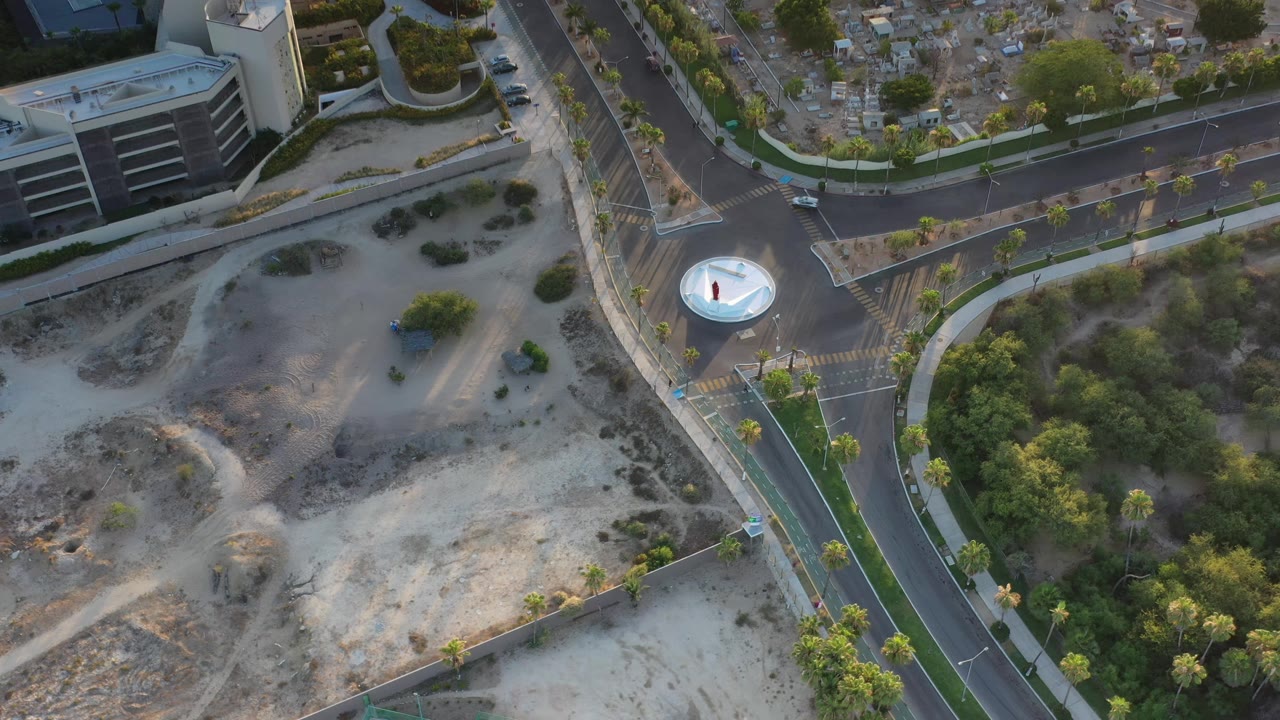 Aerial view in Los Cabos looking down on the same intersection, with a car near the upper road, green tree cover, sandy terrain, and no visible people.