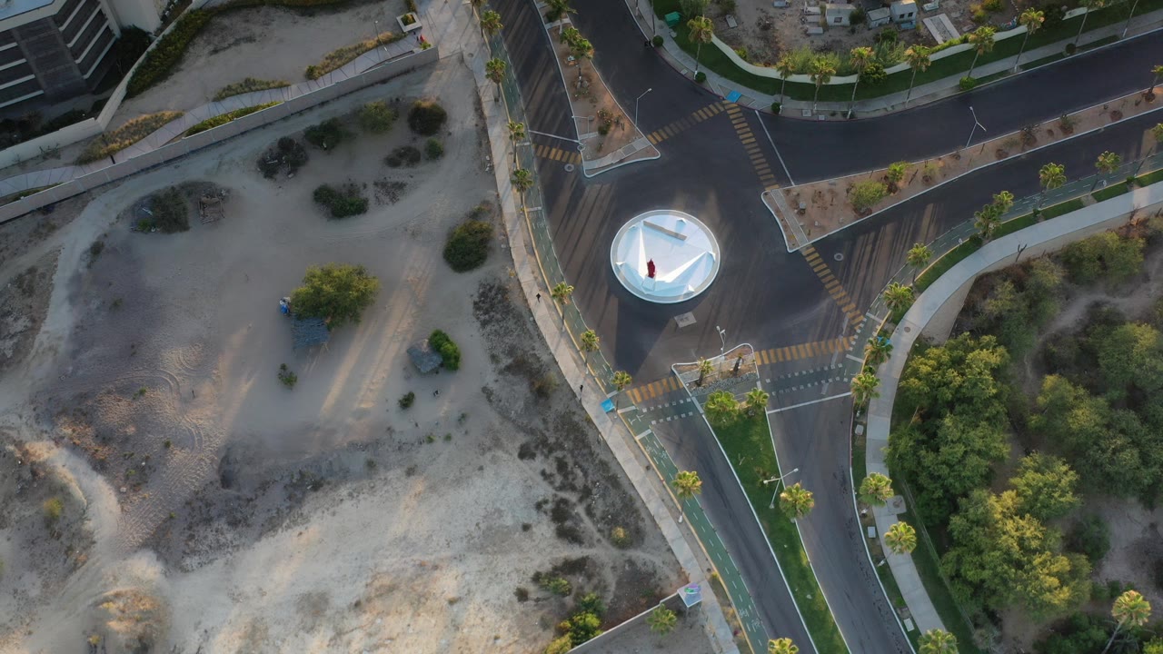 Overhead frame in Los Cabos, Mexico showing curved asphalt roads, a white circular roundabout, sidewalks, sandy lots, palms, and no visible people.