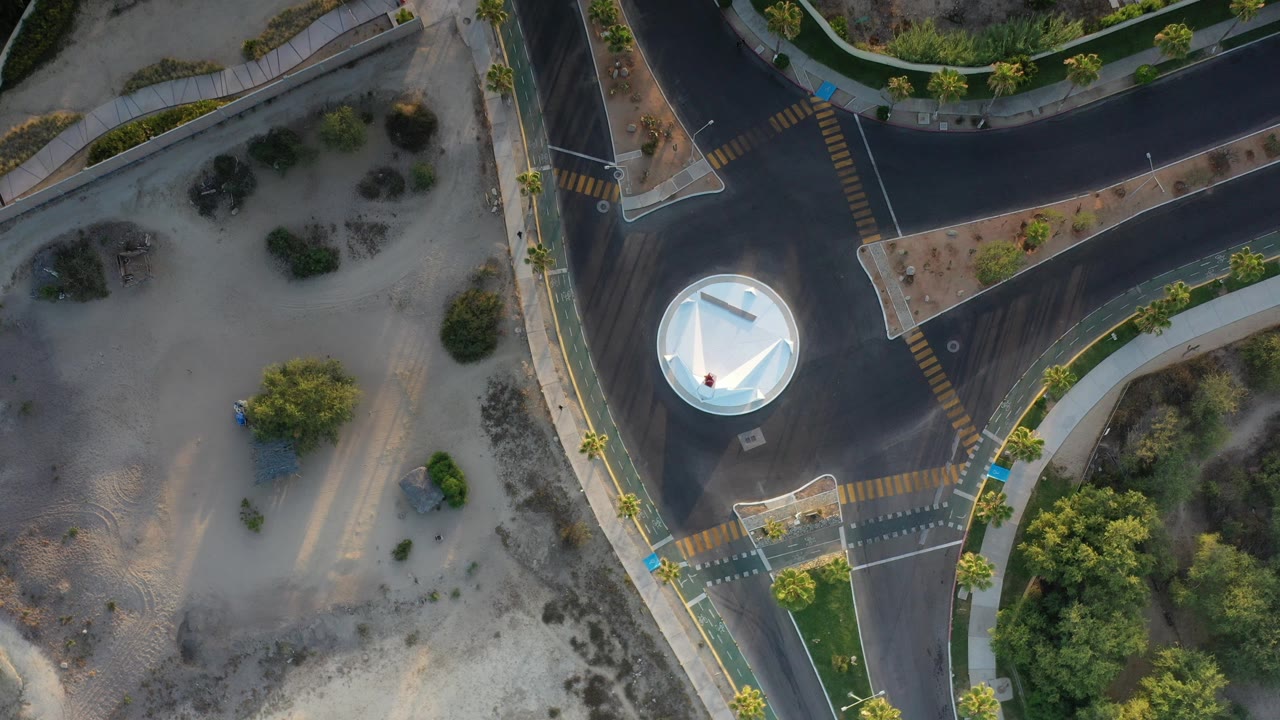 Overhead view in Los Cabos, Mexico, showing a roundabout with a white circular center, curved roads, crosswalk markings, sandy open ground, trees, and no visible people.