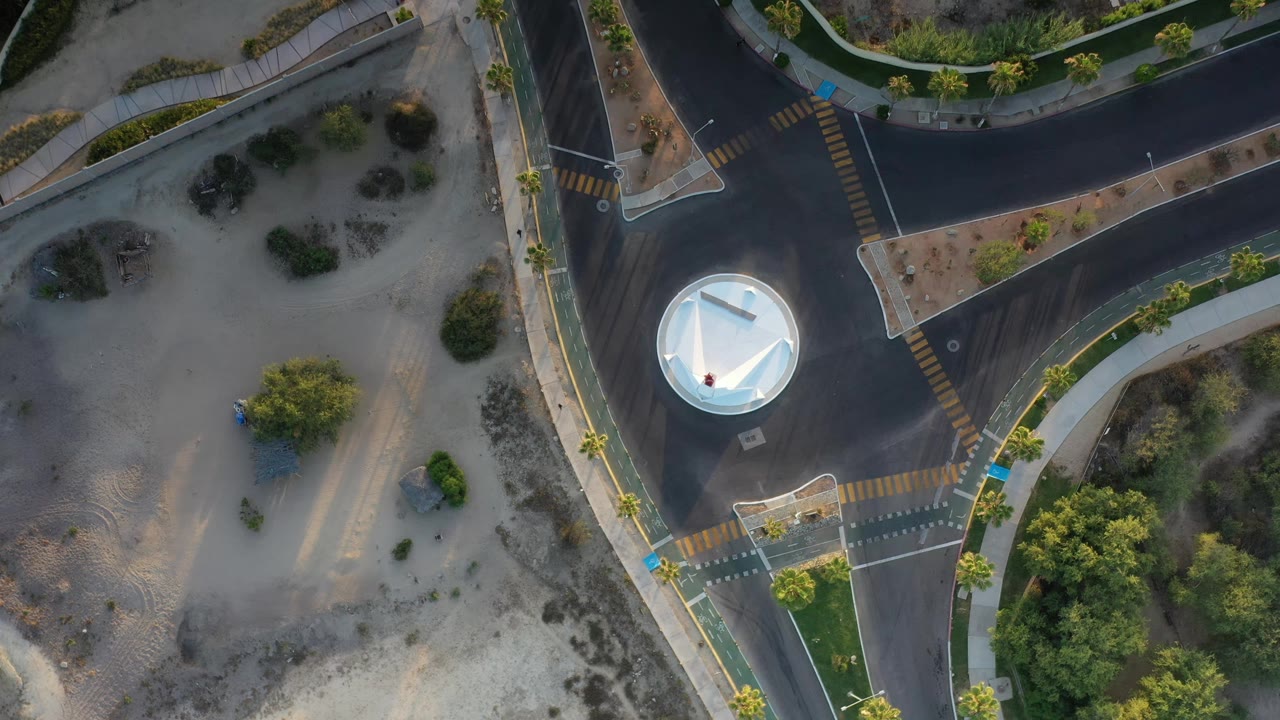 Aerial frame in Los Cabos, Mexico, with the roundabout shifted lower in view, black roads curving around landscaped medians, sandy lots, crosswalks, and no visible people.