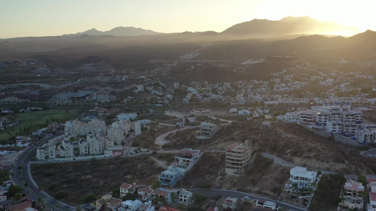 Los Cabos, Mexico appears from above as yellow sunset light flares over the mountain ridge, with clustered buildings, a curved road, dry hills, and no visible people.