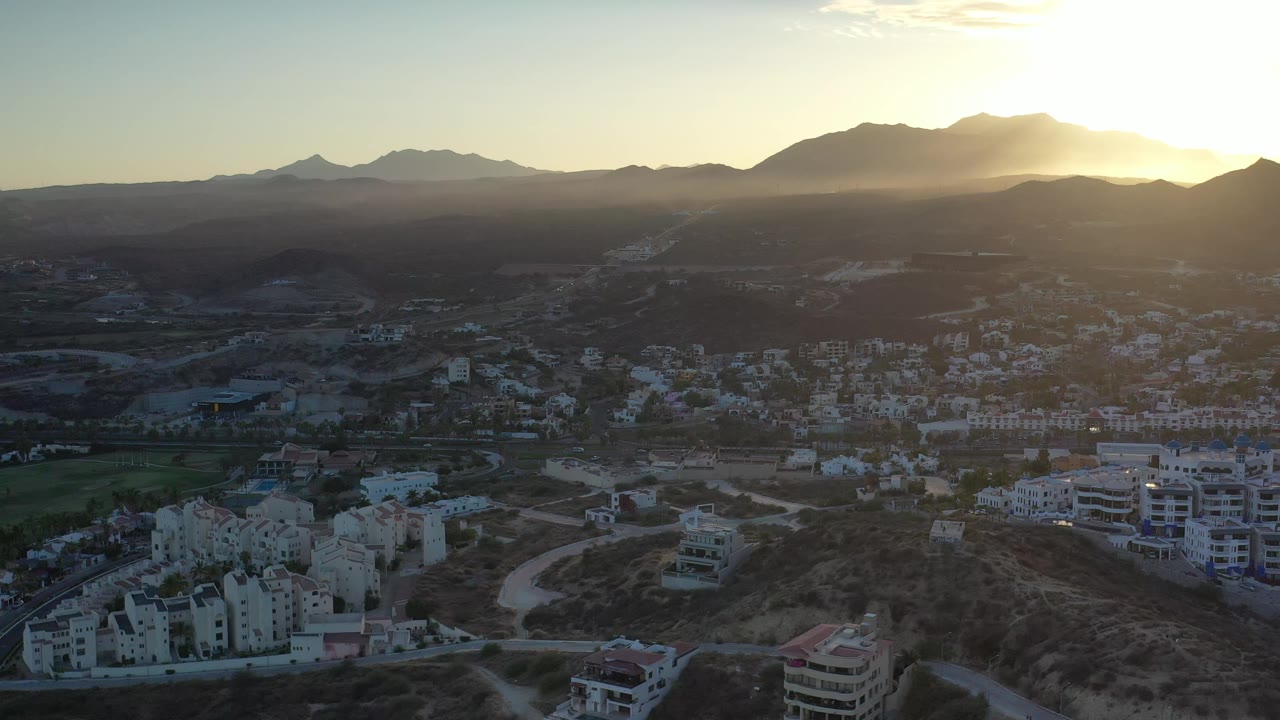 Aerial Los Cabos view with clustered white buildings in the lower frame, winding roads through dry hills, mountains across the horizon, strong sun flare, and no visible people.