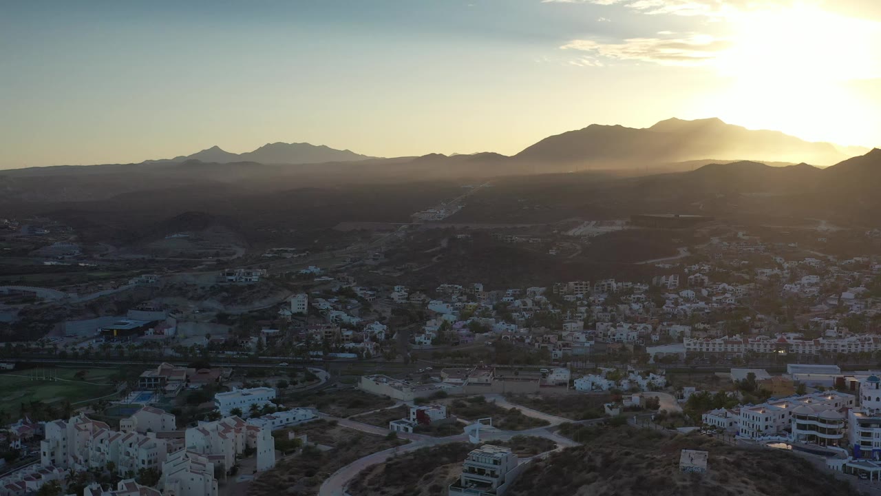 Los Cabos residential buildings sit below a broad mountain horizon, with golden light entering from the upper right and no visible people.