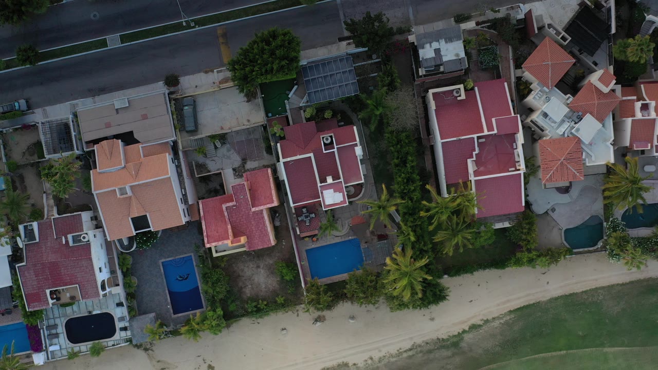 Top-down aerial frame from Los Cabos, Mexico showing red and terracotta residential roofs, small pools, palm trees, a road, and a sandy path, with no visible people.