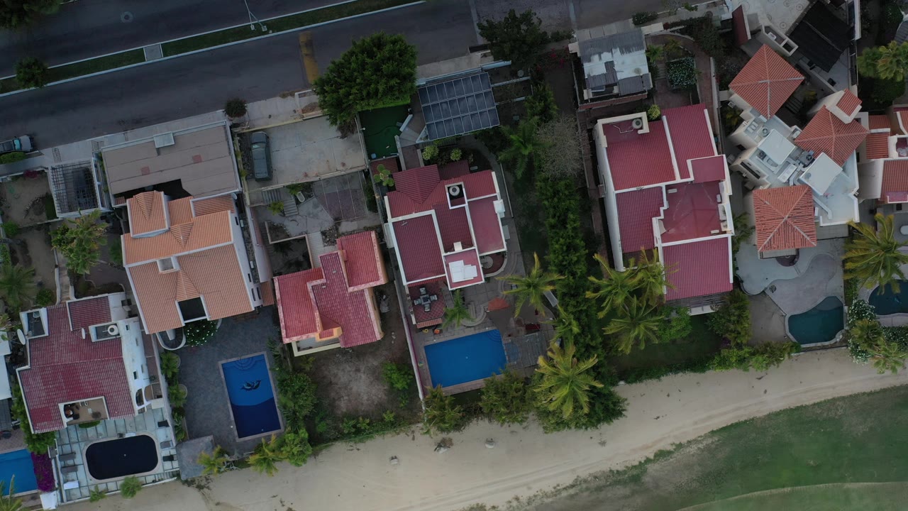 Vertical drone view of red roofs, white buildings, pools, palms, and nearby roadway.
