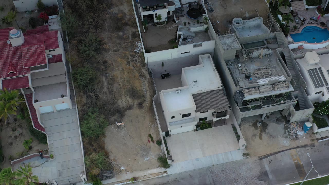 Aerial frame over Los Cabos homes with a pale driveway in the lower frame, dry hillside brush at left, red roof sections, and no visible people.