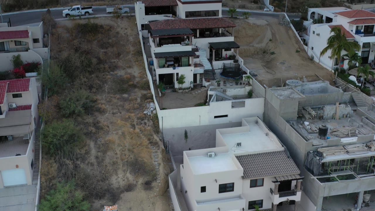 Second frame shows Los Cabos homes closer in the foreground, with a dry slope on the left, a dirt road through the center, and no visible people.