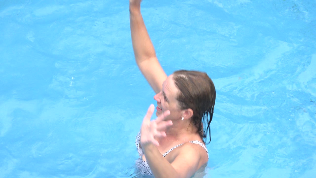 Woman in a white patterned swimsuit raises one arm in a blue swimming pool.