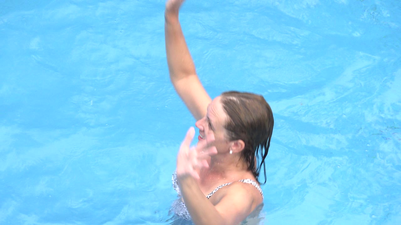 One second into the Los Cabos pool clip, the woman remains chest-deep in blue water with her arm extended upward and her other hand near her face.