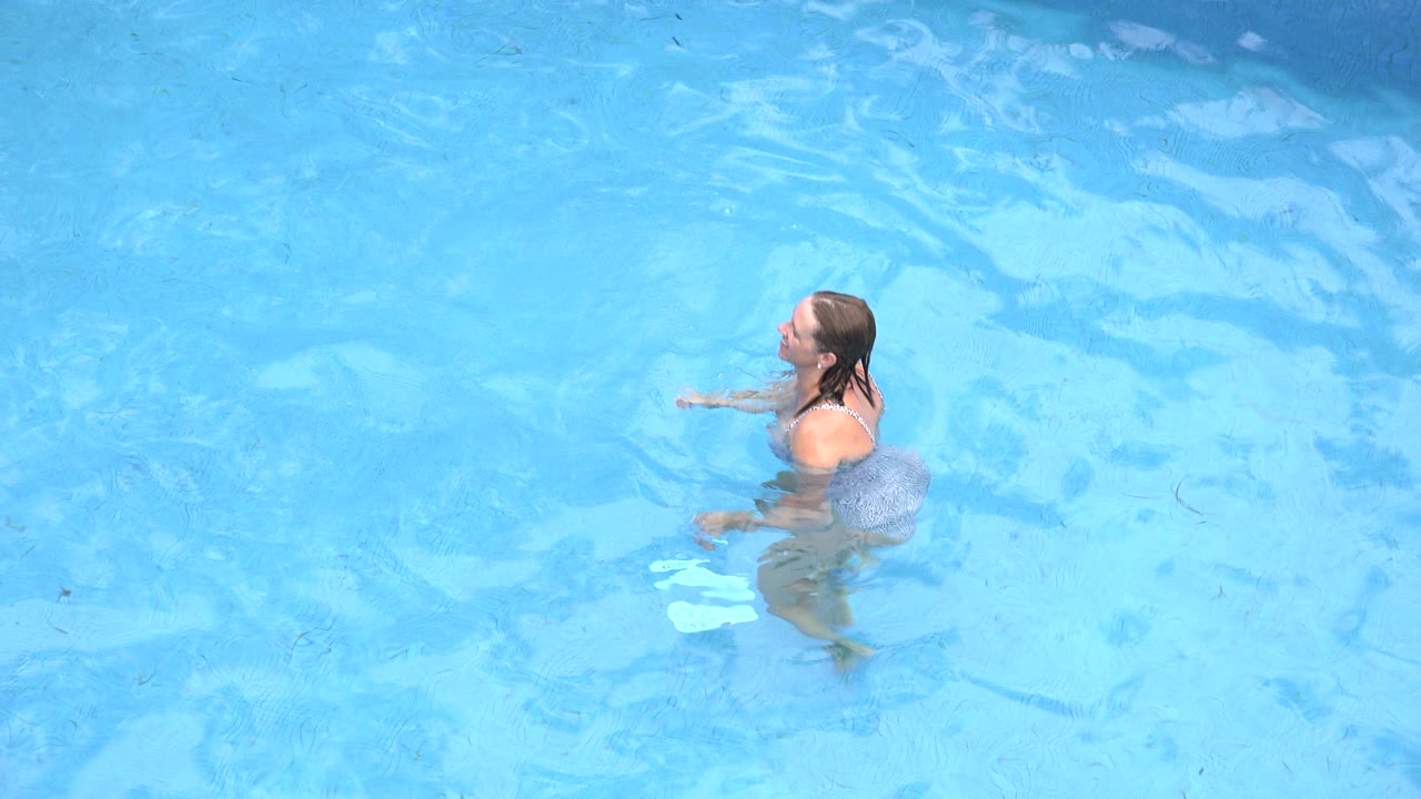 The woman remains upright in the pool, turned slightly to the side as sunlight creates pale patterns across the blue water.